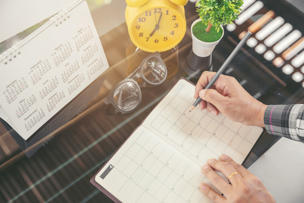Woman's hand planner or organizer writing daily appointment, mark, and noted schedule (meeting) on timetable or diary at office desk. Calendar, agenda, book, glasses, and clock on table. Planner concept